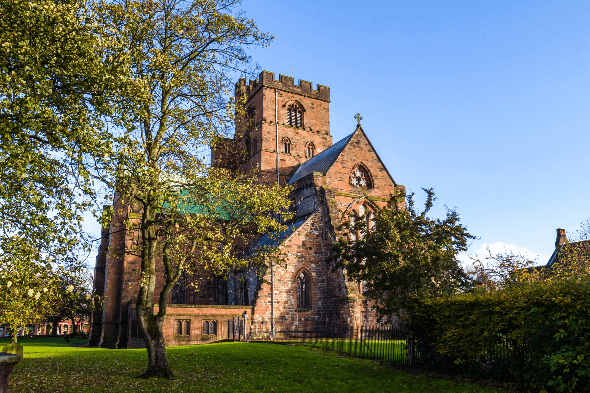 Carlisle Cathedral with trees and grass in the foreground and a bright blue sky behind