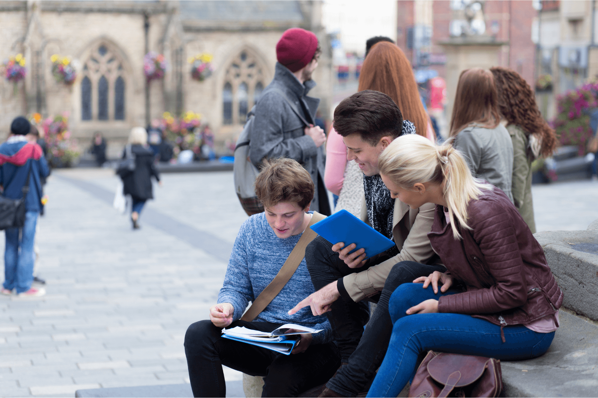 Three students sitting on stone steps looking at notes in a busy city centre with other students in the background nearby