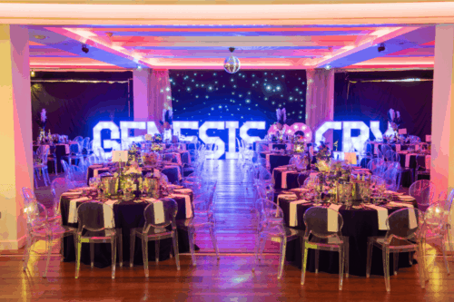 Large lit up letters spelling GENESIS CRY in the background behind dressed tables ready for the event