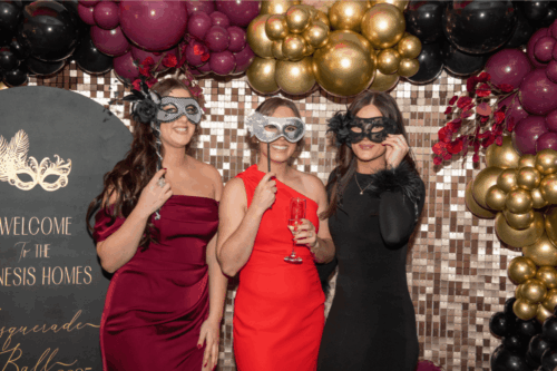 Three women in ball attire holding masquerade masks over their faces in front of metallic backdrop decorations