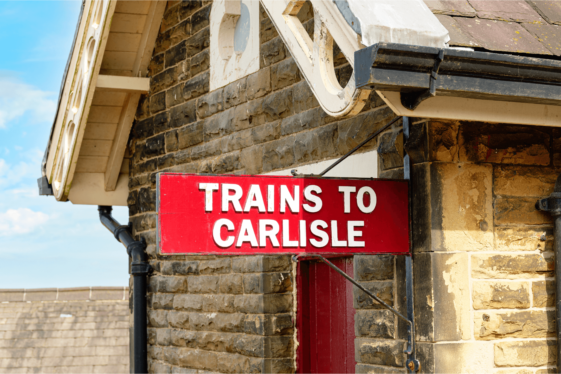 Close up of a red sign on a stone train station wall saying 'trains to Carlisle'