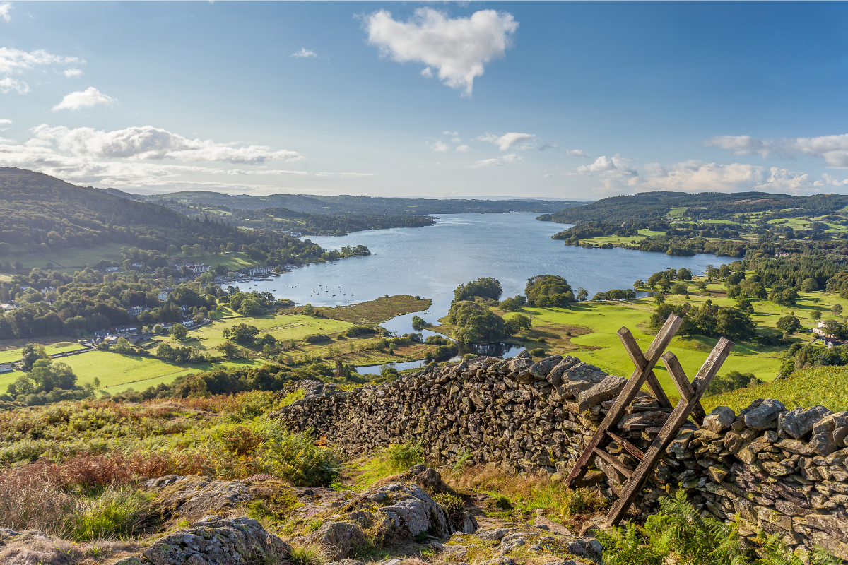 View of Windermere in the English Lake District taken from Loughrigg Fell. On a summer morning. With white fluffy clouds, shafts of Light on to Green fields with a Dry Stone Wall and wooden Style in the Foreground.